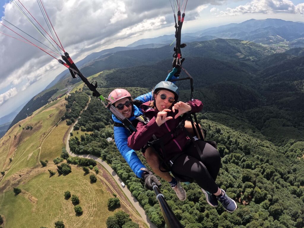 Photo prise en plein vol en parapente biplace, avec une passagère qui fait un coeur avec ses mains. La photo est prise par le pilote